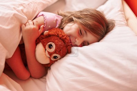 Photo for Adorable hispanic girl hugging monkey doll lying on bed at bedroom - Royalty Free Image