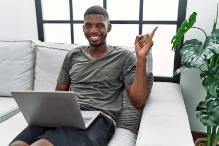 Photo for Young african american man using laptop at home sitting on the sofa with a big smile on face, pointing with hand finger to the side looking at the camera. - Royalty Free Image