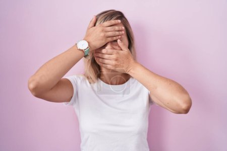 Photo for Young blonde woman standing over pink background covering eyes and mouth with hands, surprised and shocked. hiding emotion - Royalty Free Image