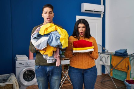 Foto de Young couple holding laundry dirty and clean laundry in shock face, looking skeptical and sarcastic, surprised with open mouth - Imagen libre de derechos