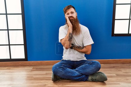 Téléchargez les photos : Homme rousse à longue barbe assis sur le sol dans une pièce vide pensant fatigué et ennuyé par des problèmes de dépression avec les bras croisés. - en image libre de droit