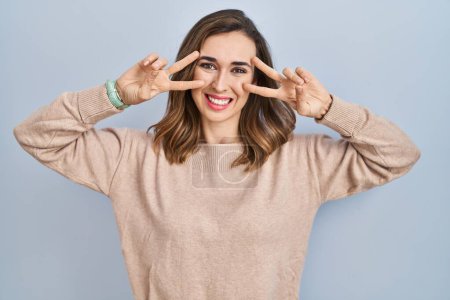 Photo for Young woman standing over isolated background doing peace symbol with fingers over face, smiling cheerful showing victory - Royalty Free Image