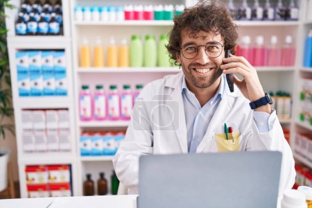 Photo for Young hispanic man pharmacist talking on smartphone using laptop at pharmacy - Royalty Free Image