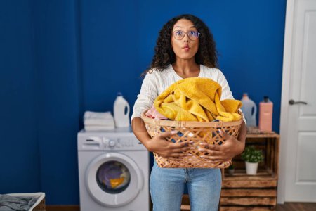 Photo for Young hispanic woman doing laundry holding wicker basket making fish face with mouth and squinting eyes, crazy and comical. - Royalty Free Image