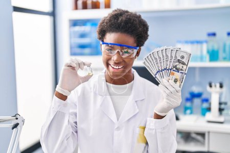 Foto de Mujer afroamericana trabajando en laboratorio científico sosteniendo dólares sonriendo y riendo en voz alta porque divertida broma loca. - Imagen libre de derechos