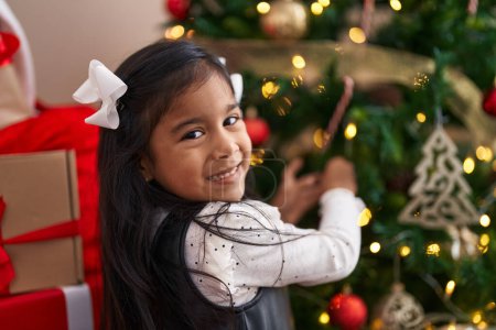 Foto de Adorable chica hispana sonriendo confiada decorando el árbol de Navidad en casa - Imagen libre de derechos