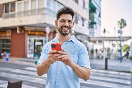 Foto de Joven hombre hispano sonriendo feliz usando smartphone en la ciudad. - Imagen libre de derechos