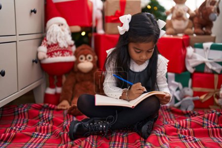 Photo for Adorable hispanic girl writing on notebook sitting on floor by christmas tree at home - Royalty Free Image