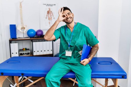 Photo for Young physiotherapist man working at pain recovery clinic doing ok gesture with hand smiling, eye looking through fingers with happy face. - Royalty Free Image