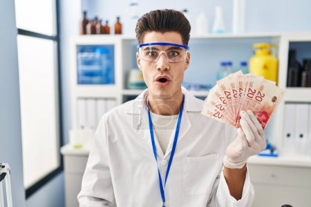 Photo for Young hispanic man working at scientist laboratory holding shekels scared and amazed with open mouth for surprise, disbelief face - Royalty Free Image