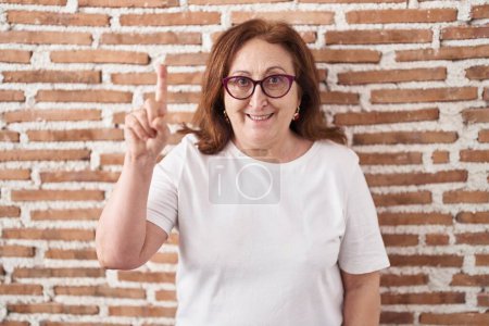 Foto de Mujer mayor con gafas de pie sobre la pared de ladrillos que muestra y señala hacia arriba con el dedo número uno mientras sonríe confiado y feliz. - Imagen libre de derechos