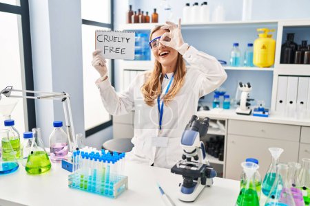 Photo for Young blonde scientist woman working on cruelty free laboratory smiling happy doing ok sign with hand on eye looking through fingers - Royalty Free Image