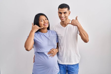 Photo for Young hispanic couple expecting a baby standing over background smiling doing phone gesture with hand and fingers like talking on the telephone. communicating concepts. - Royalty Free Image