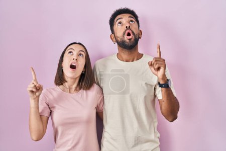 Téléchargez les photos : Young hispanic couple together over pink background amazed and surprised looking up and pointing with fingers and raised arms. - en image libre de droit