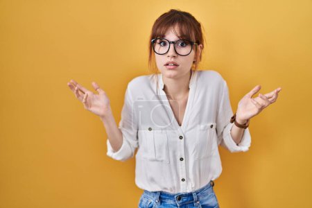Foto de Young beautiful woman wearing casual shirt over yellow background clueless and confused expression with arms and hands raised. doubt concept. - Imagen libre de derechos