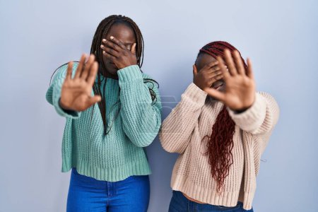 Photo for Two african woman standing over blue background covering eyes with hands and doing stop gesture with sad and fear expression. embarrassed and negative concept. - Royalty Free Image