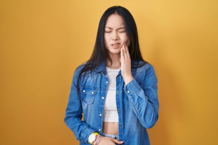 Foto de Young asian woman standing over yellow background touching mouth with hand with painful expression because of toothache or dental illness on teeth. dentist - Imagen libre de derechos