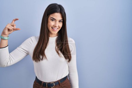 Photo for Young brunette woman standing over blue background smiling and confident gesturing with hand doing small size sign with fingers looking and the camera. measure concept. - Royalty Free Image