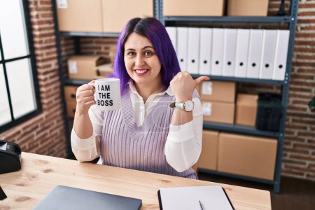 Foto de Plus size woman wit purple hair working at small business ecommerce holding i am the boss cup pointing thumb up to the side smiling happy with open mouth - Imagen libre de derechos