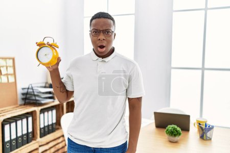Foto de Hombre africano joven sosteniendo el despertador en la oficina asustado y sorprendido con la boca abierta para sorpresa, cara de incredulidad - Imagen libre de derechos