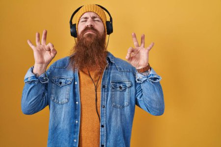 Téléchargez les photos : Homme caucasien à longue barbe écoutant de la musique à l'aide d'écouteurs détendus et souriants avec les yeux fermés faisant geste de méditation avec les doigts. concept de yoga. - en image libre de droit
