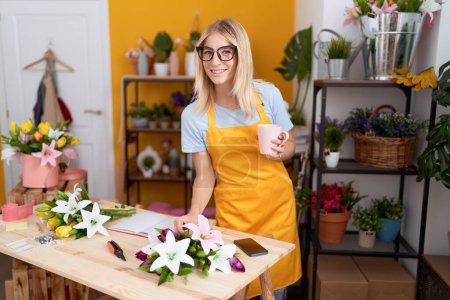 Téléchargez les photos : Jeune femme blonde fleuriste boire une tasse de café écrit sur ordinateur portable à la boutique de fleurs - en image libre de droit
