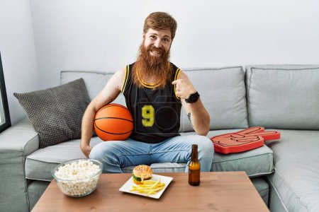 Foto de Hombre caucásico con barba larga sosteniendo pelota de baloncesto animando juego de tv mirando confiado con sonrisa en la cara, señalándose con los dedos orgullosos y felices. - Imagen libre de derechos