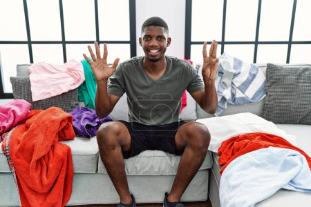 Foto de Joven afroamericano sentado en el sofá con ropa sucia mostrando y apuntando hacia arriba con los dedos número ocho mientras sonríe confiado y feliz. - Imagen libre de derechos