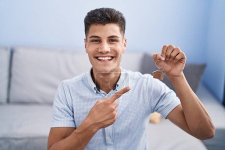 Foto de Joven hombre hispano sosteniendo las llaves de un nuevo hogar sonriendo feliz señalando con la mano y el dedo - Imagen libre de derechos