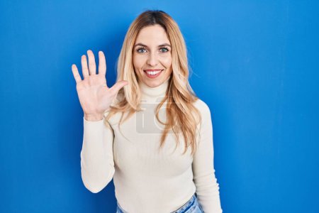 Téléchargez les photos : Jeune femme caucasienne debout sur fond bleu montrant et pointant vers le haut avec les doigts numéro cinq tout en souriant confiant et heureux. - en image libre de droit