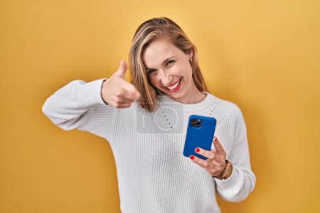 Téléchargez les photos : Jeune femme blonde à l'aide d'un smartphone tapant message pointant des doigts vers la caméra avec un visage heureux et drôle. bonne énergie et vibes. - en image libre de droit