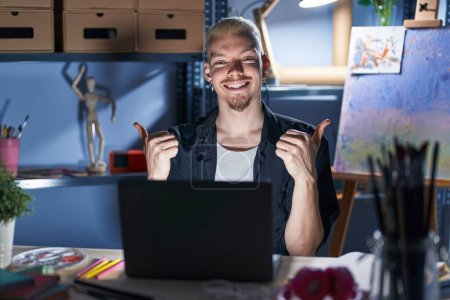Photo for Young caucasian man using laptop at night at art studio success sign doing positive gesture with hand, thumbs up smiling and happy. cheerful expression and winner gesture. - Royalty Free Image