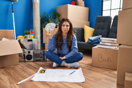 Photo for Young brunette woman sitting on the floor at new home holding money thinking attitude and sober expression looking self confident - Royalty Free Image