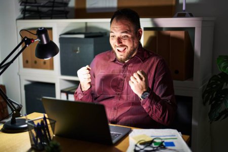 Foto de Plus size hispanic man with beard working at the office at night celebrating surprised and amazed for success with arms raised and open eyes. winner concept. - Imagen libre de derechos