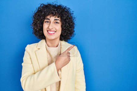 Foto de Young brunette woman with curly hair standing over blue background cheerful with a smile on face pointing with hand and finger up to the side with happy and natural expression - Imagen libre de derechos