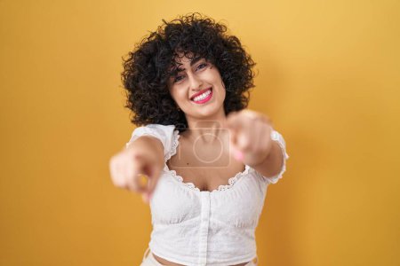 Foto de Young brunette woman with curly hair standing over yellow background pointing to you and the camera with fingers, smiling positive and cheerful - Imagen libre de derechos