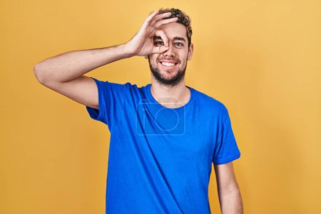 Foto de Hombre hispano con barba de pie sobre fondo amarillo haciendo buen gesto con la mano sonriente, ojos mirando a través de los dedos con cara feliz. - Imagen libre de derechos