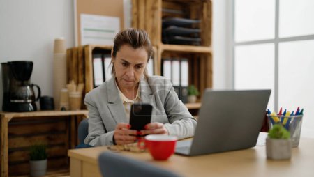 Foto de Trabajadora de negocios hispana de mediana edad usando teléfono inteligente en la oficina - Imagen libre de derechos