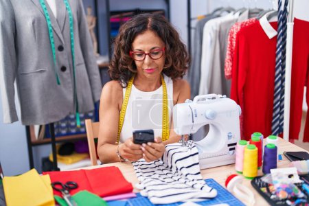 Téléchargez les photos : Femme d'âge moyen tailleur en utilisant smartphone à l'usine de vêtements - en image libre de droit