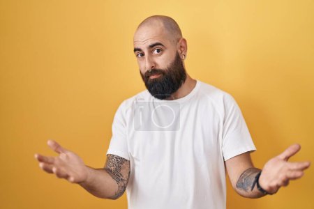 Foto de Young hispanic man with beard and tattoos standing over yellow background clueless and confused with open arms, no idea concept. - Imagen libre de derechos