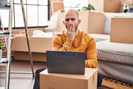 Photo for Young bald man with beard moving to a new house using laptop covering mouth with hand, shocked and afraid for mistake. surprised expression - Royalty Free Image