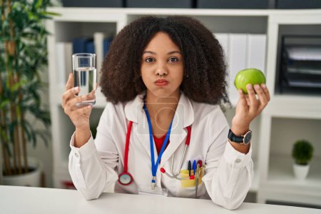 Foto de Young african american dietitian woman holding fresh apple and water puffing cheeks with funny face. mouth inflated with air, catching air. - Imagen libre de derechos