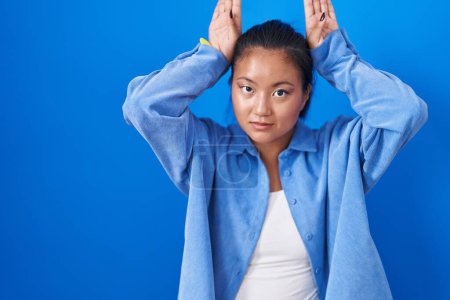 Foto de Asian young woman standing over blue background doing bunny ears gesture with hands palms looking cynical and skeptical. easter rabbit concept. - Imagen libre de derechos