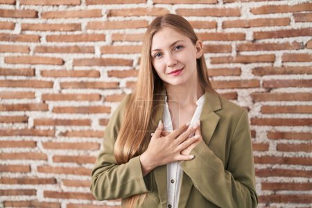 Téléchargez les photos : Jeune femme caucasienne debout sur le mur de briques fond souriant avec les mains sur la poitrine avec les yeux fermés et geste reconnaissant sur le visage. concept de santé. - en image libre de droit