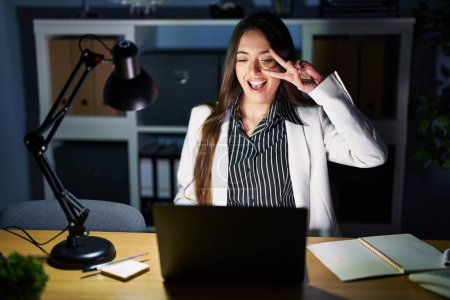 Foto de Mujer morena joven que trabaja en la oficina por la noche con el ordenador portátil haciendo símbolo de la paz con los dedos en la cara, sonriendo alegre mostrando la victoria - Imagen libre de derechos