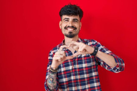 Téléchargez les photos : Jeune homme hispanique avec la barbe debout sur fond rouge souriant dans l'amour faisant forme de symbole de coeur avec les mains. concept romantique. - en image libre de droit