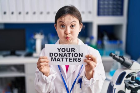 Photo for Woman with down syndrome working at scientist laboratory holding your donation matters banner puffing cheeks with funny face. mouth inflated with air, catching air. - Royalty Free Image