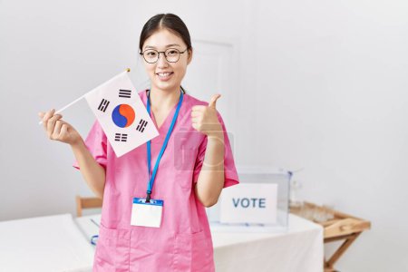 Téléchargez les photos : Jeune infirmière asiatique femme à l'élection de la campagne politique tenant drapeau de la Corée du Sud souriant heureux et positif, pouce levé faire excellent et signe d'approbation - en image libre de droit