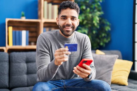 Foto de Joven hombre hispano usando teléfono inteligente y tarjeta de crédito sentado en el sofá en casa - Imagen libre de derechos
