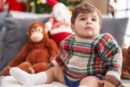 Photo for Adorable hispanic toddler sitting on sofa by christmas tree at home - Royalty Free Image